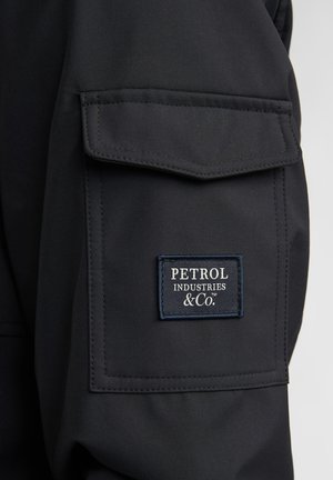 Giacca nera con tasca frontale dotata di etichetta rettangolare in tessuto. L'etichetta presenta testo bianco: "PETROL INDUSTRIES & Co." su sfondo blu scuro.
