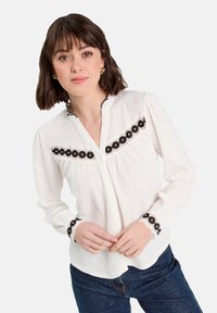 Blouse blanche texturée avec une bordure en dentelle florale noire le long de l'encolure et des poignets, présentant une fermeture à boutons sur le devant et des manches longues.