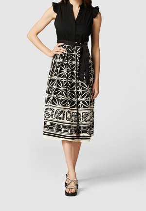 Kobieta nosząca czarną sukienkę bez rękawów z czarno-białą geometryczną midi spódnicą i czarnymi sandałami na paski.