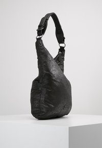 Musta keinonahkainen olkalaukku, jossa on rypistynyt pinta, epäsymmetrinen muoto, säädettävä hihna ja vetoketjukiinnitys.