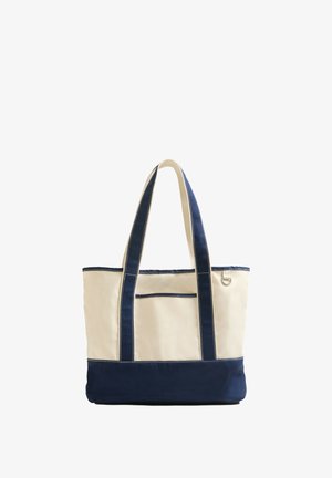 Plátěná taška s krémovým vrškem, navy modrým spodkem a odpovídajícími uchy. Obsahuje přední kapsu a kontrastní stehy.