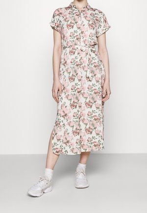 Kvinde iført en midi-kjole med korte ærmer, bælte, knapper og pink blomsterprint samt hvide sneakers foran en ensfarvet baggrund.