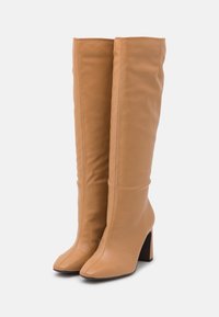 Bottes en cuir beige montantes avec une texture lisse, à bout carré et à talon épais. Coutures visibles le long de la tige.
