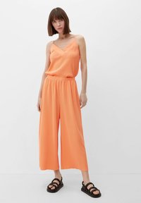 Oranje mouwloos jumpsuit met een v-hals, elastische tailleband en wijde broekspijpen. Gecombineerd met zwarte gekruiste sandalen. Gladde stofstructuur.