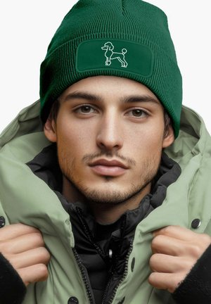 Green knit beanie with a patch featuring a dog outline. Model wears a light green puffy jacket with a black inner layer, capturing close-up.