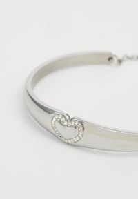 Bracciale tono argento caratterizzato da un design elegante e lucido con un accento a forma di cuore decorato con strass trasparenti. Texture liscia su tutta la superficie.