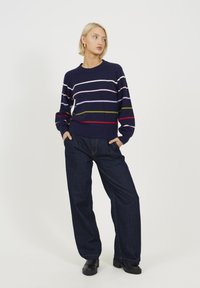 Sweter w niebieskie paski z lawendowymi, zielonymi i czerwonymi liniami; zestawiony z luźnymi ciemnymi dżinsami. Model stoi z rękami w kieszeniach.