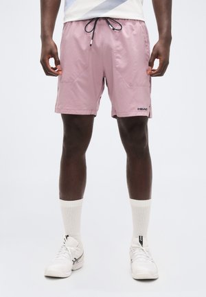 Άνδρας που φοράει ροζ αθλητικό σορτς με κορδόνι, λευκές κάλτσες μέχρι τον αστράγαλο και λευκά αθλητικά παπούτσια, στέκεται μπροστά σε απλό φόντο.