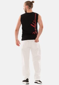 Schwarzes ärmelloses Shirt mit rotem Schriftzug auf dem Rücken, kombiniert mit weißen, locker sitzenden Hosen und schwarzen Sneakern. Einfacher, lässiger Stil.