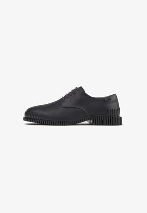 Zapato de vestir de cuero negro con suela gruesa ranurada y cordones, visto de lado sobre un fondo blanco.