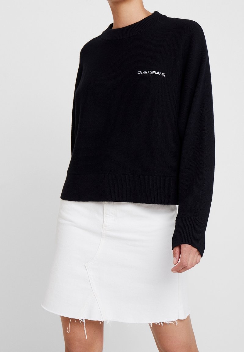 Czarny dzianinowy sweter z okrągłym dekoltem i ściągaczami, z białym logo "Calvin Klein Jeans" na piersi, noszony z białą spódnicą dżinsową.