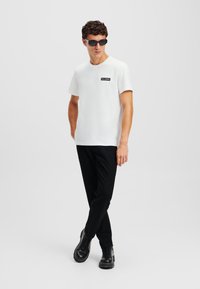 Cămașă albă cu mânecă scurtă, dotată cu un petic de logo negru, asortată cu pantaloni negri și bocanci negri cu șireturi, pe un fundal neutru.