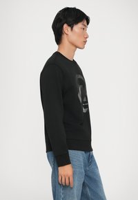 Μαύρη μπλούζα με textured γραφικό σχέδιο που απεικονίζει ένα πρόσωπο, στρογγυλή λαιμόκοψη και ριπτές μανσέτες. Συνδυασμένη με ανοιχτό μπλε τζιν.