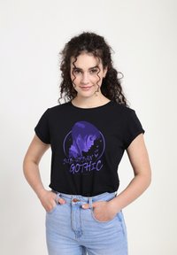Mladá žena s kudrnatými vlasy, oblékající černé tričko "Sub-Urban Gothic" a světle modré džíny, stojící s rukama v kapsách na bílém pozadí.