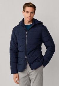 Chaqueta acolchada azul marino con capucha, con cremallera frontal, bolsillos laterales y diseño acolchado. Textura suave con un corte moderno.