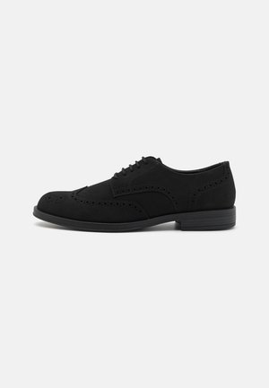 Chaussure Oxford en cuir noir à bout golf, avec des détails brogue et une fermeture à lacets, présentée sur un fond clair uni.