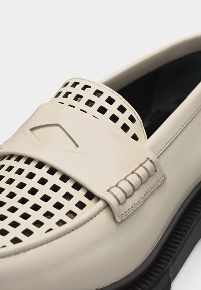 Κρεμ μοκασίνι με διάτρητο τετραγωνικό σχέδιο, ραμμένη ταινία στην κορυφή και μαύρη σόλα, εμφανής σε λευκό φόντο.