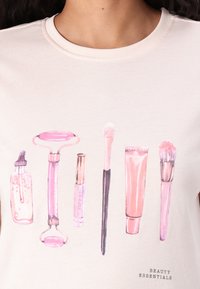 Crème t-shirt met een grafisch ontwerp van schoonheidsmiddelen in het roze, waaronder een roller, kwast en flessen, met de tekst "SCHOONHEIDS BASISPRODUCTEN" eronder.