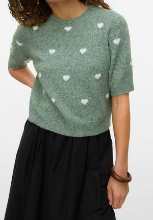 Femme portant un pull vert à manches courtes avec des motifs de cœurs blancs et une jupe noire, tenant un bracelet marron à son poignet.