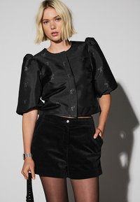 Melna puskrekla ar pūkainām piedurknēm, kurā ir sudraba loku izšuvums. Saskaņota ar melnā krāsā korduroja šortiem un melnām zeķubiksēm.