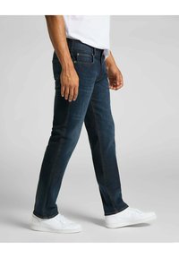 Homme portant un jean bleu foncé et des baskets blanches, debout avec une jambe légèrement en avant sur un fond gris clair.