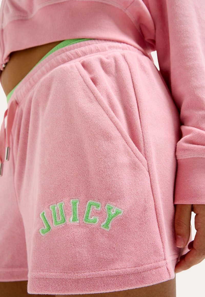 Personne portant un short en tissu éponge rose et un haut à manches longues assorti, avec une ceinture verte et "JUICY" brodé en vert sur le short.