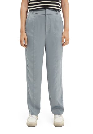 Persoon die lichtblauwe satijnen broek met rechte pijpen draagt, witte sneakers en een zwart-wit gestreept mouwloos topje.