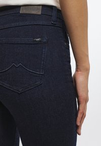 Mörkblå denimjeans med en åtsittande design, med en läderlapp vid midjan och en karaktäristisk böjd söm vid fickan.