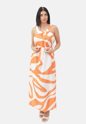 Kvinde med glat mørkt hår iført en hvid og orange ærmeløs maxikjole med abstrakte mønstre, der binder et taljebælte, stående mod en hvid baggrund.