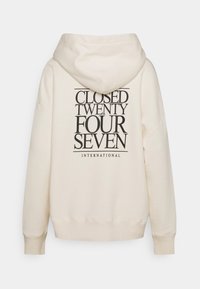 Sweat à capuche crème avec un grand texte noir sur le dos indiquant "CLOSED TWENTY FOUR SEVEN INTERNATIONAL", avec une capuche à cordon.