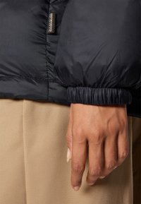 Main dans la poche d'un pantalon beige, portant une doudoune noire avec un poignet élastique et une étiquette Napapijri visible.