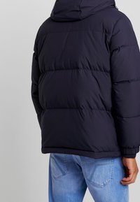 Person, der eine dunkle gepolsterte Kapuzenjacke und hellblaue Jeans trägt, von hinten vor einem schlichten hellen Hintergrund betrachtet.