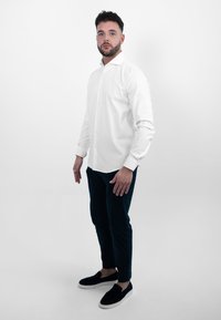 Witte button-up blouse met lange mouwen, gecombineerd met donkerblauwe broek en zwarte instapschoenen. Glad materiaal en getailleerde pasvorm.