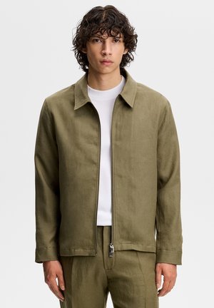 Młody mężczyzna o kręconych włosach, ubrany w oliwkowo-zieloną kurtkę na zamek z białym T-shirtem, stojący na tle jednolitego tła.