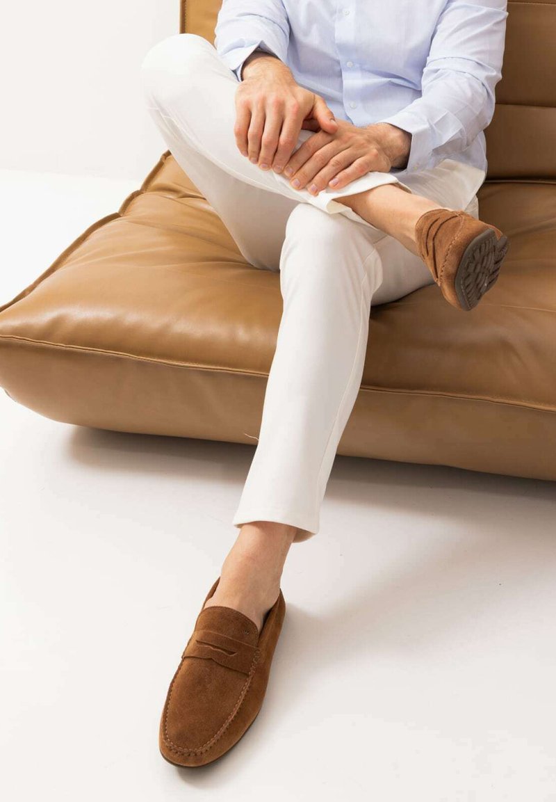 Braune Wildleder-Slipper mit gesticktem Design und flacher Gummisohle, getragen mit weißen Hosen, auf einem beigefarbenen Ledersofa sitzend.