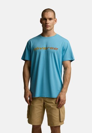 Jasnoniebieska bawełniana koszulka z krótkimi rękawami, z napisem "różnorodny" w brązowych literach na klatce piersiowej. Noszona z beżowymi spodniami cargo.