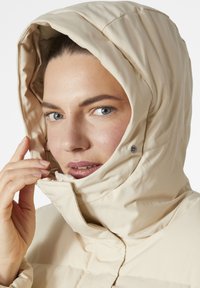 Béžová prošívaná bunda s vysokým límcem a připevněnou kapucí. Tkanina se zdá být hladká a poblíž brady jsou patentky.