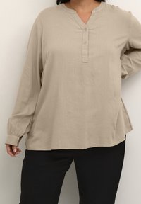 Beżowa bluzka z długim rękawem, zaokrąglonym kołnierzem, plisą na guziki i luźnym krojem; wykonana z lekkiego materiału o gładkiej fakturze.