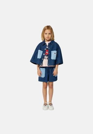 Denimoutfit met een cropped jack met blauwe patchzakken, gecombineerd met bijpassende shorts. Witte sneakers met tijgerborduursel maken de look compleet.
