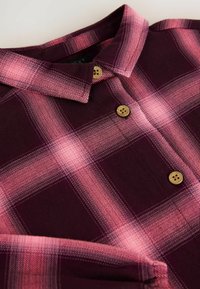 Camicia a quadri in tonalità di borgogna scuro e rosa, con bottoni in legno, colletto e una texture morbida. Cuciture e pieghe visibili.