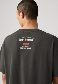 Camiseta de algodón gris con un estampado en la parte posterior que incluye "Toy Story", "Levi's" y "Ropa Occidental Auténtica". Mangas cortas y ajuste relajado.