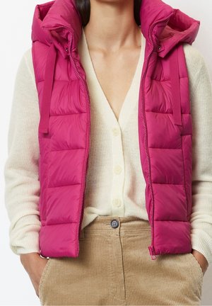 Kvinde iført en lyserød, quiltet, ærmeløs vest over en cremefarvet, knappet cardigan og beige kordfløjlsbukser, hænderne i lommerne.