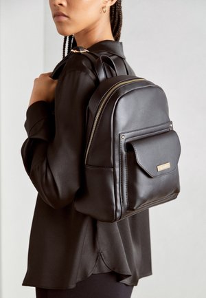 Kobieta nosząca czarny skórzany plecak z złotym zamkiem i przednią kieszenią, ubrana w czarną koszulę z długim rękawem, na jednolitym tle.