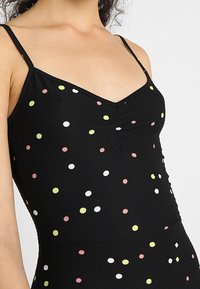 Czarny bezrękawnik z cienkimi ramiączkami, wykonany z teksturowanej tkaniny, pokryty kolorowymi kropeczkami w odcieniach różu, żółci i bieli. Dekolt w kształcie litery V.