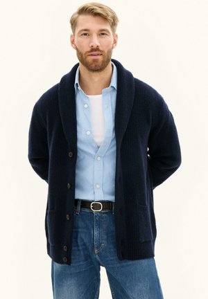Skægget mand iført en marineblå cardigan over en lyseblå skjorte med knapper og en hvid undertrøje, kombineret med blå jeans og et sort bælte.