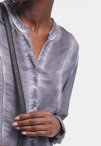 Camicia grigia a maniche lunghe realizzata in un tessuto morbido, caratterizzata da una leggera lucentezza, un colletto a fascia e cuciture a contrasto lungo la patta.
