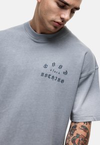 Szary, oversize bawełniany t-shirt z okrągłym dekoltem i nadrukiem tekstowym z przodu w ciemnoszarym kolorze. Krótkie rękawy, gładka tekstura, luźny krój.