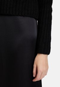Schwarzer Strickpullover aus Stoff und glatter, schwarzer Satinrock. Der Ärmel ist gerippt und der Rock hat eine elegante Textur, auf dem eine Hand ruht.
