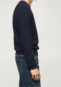 Mężczyzna ubrany w ciemnoniebieski sweter w drobne wzory i niebieskie dżinsy, stojący w profilu na tle jasnego, jednolitego tła.