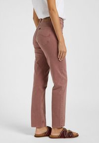 Jeans rectos de color rosa con una cintura alta, que presentan un diseño de bolsillo trasero. Llevados con sandalias marrones que muestran acentos decorativos.